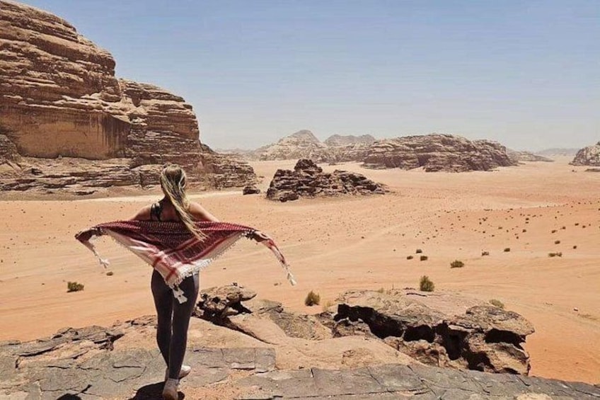 A person standing on a rocky outcrop in a desert landscape with sparse vegetation and distant mountains. 2