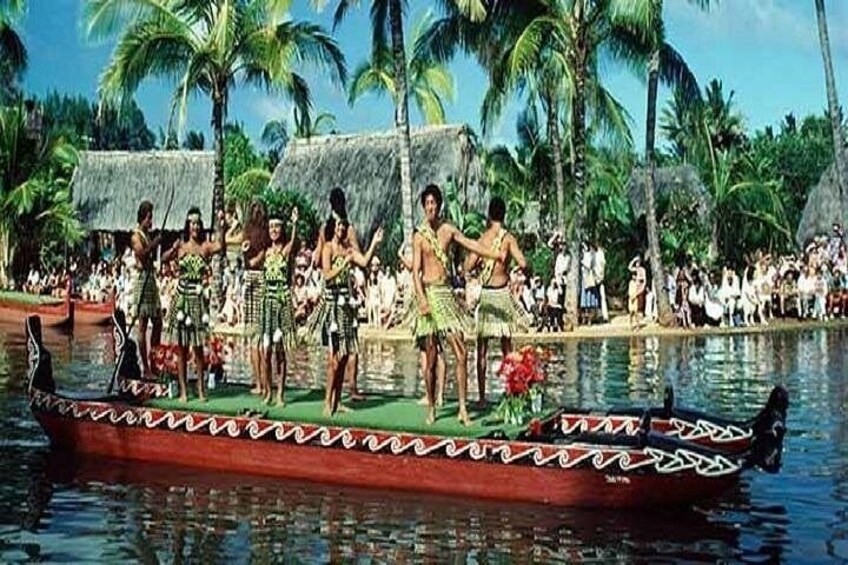 Una canoa tradicional con gente vestida de verde y amarillo, una multitud en la orilla y palmeras al fondo. 1