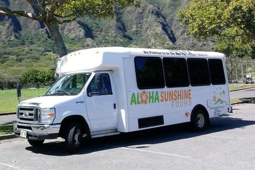 Un autobús blanco con la marca 'Aloha Sunshine Tours' estacionado en una carretera con montañas al fondo. 5