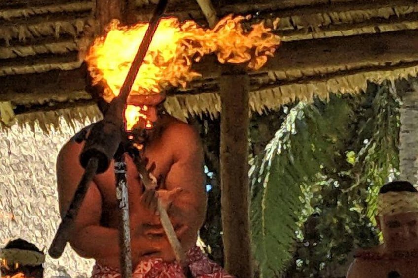 A person with a large fire on their head, wearing a traditional headdress, in a thatched-roof structure with greenery in the background. 2