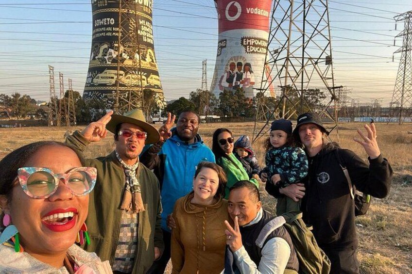 A group of people posing in front of a water tower with graffiti and a power line structure. 4