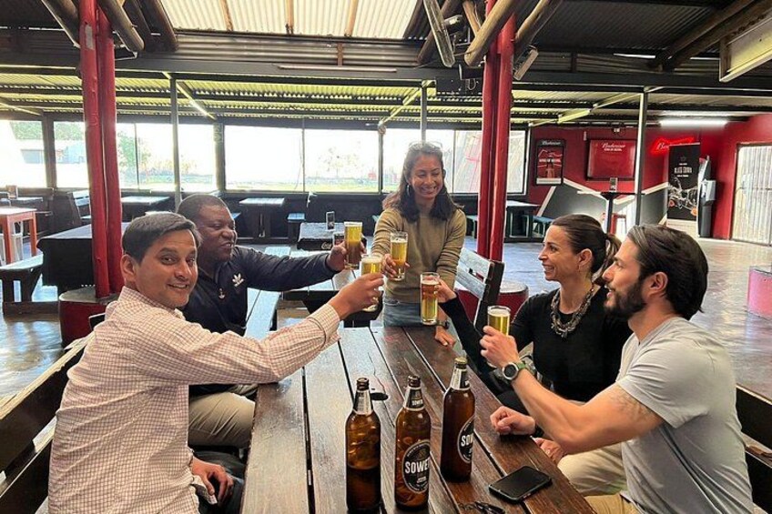 A group of people toasting with beer at a wooden table in a restaurant with red pillars and a metal roof. 2