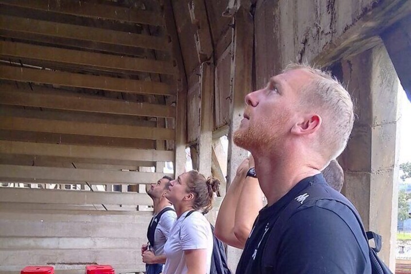A person standing under a wooden structure, possibly a bridge or shelter, with a metal railing visible in the background. 1