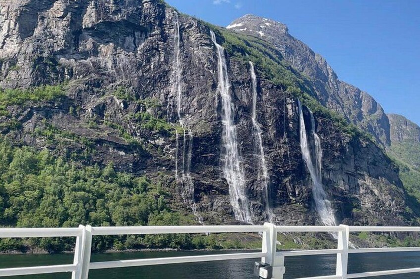 A waterfall cascading down a rocky cliff, a body of water in the foreground, and a forested hillside. 2