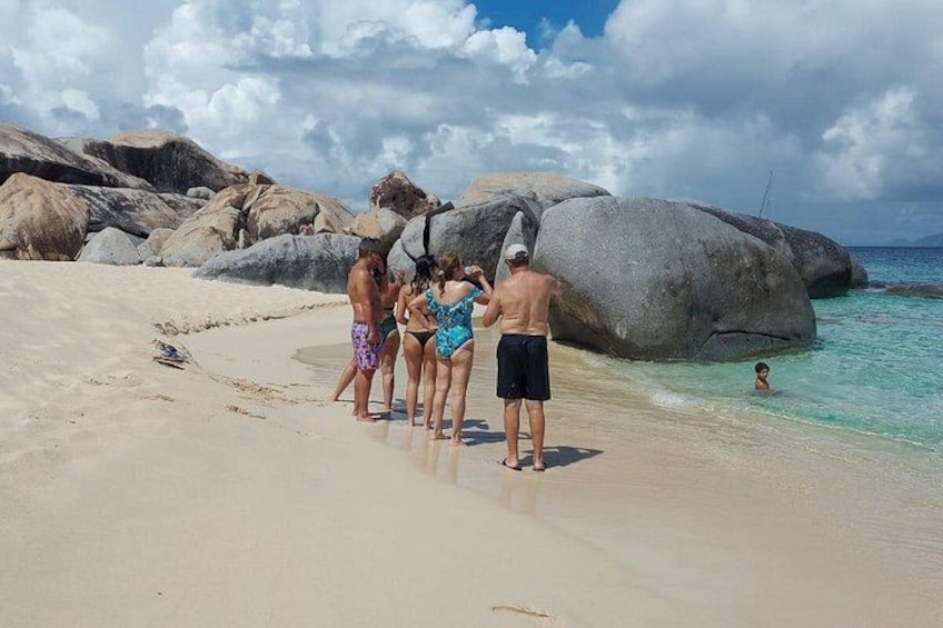 A group of people on a sandy beach with large rocks and clear blue water. 1