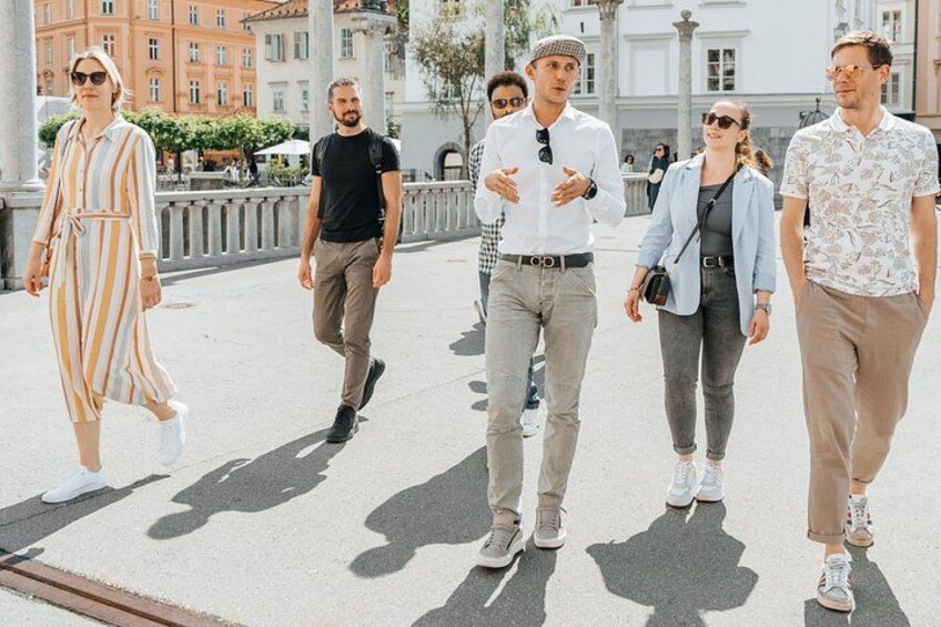 En grupp människor promenerar på en asfalterad gång i en stadsmiljö, med byggnader och en fontän i bakgrunden. 3