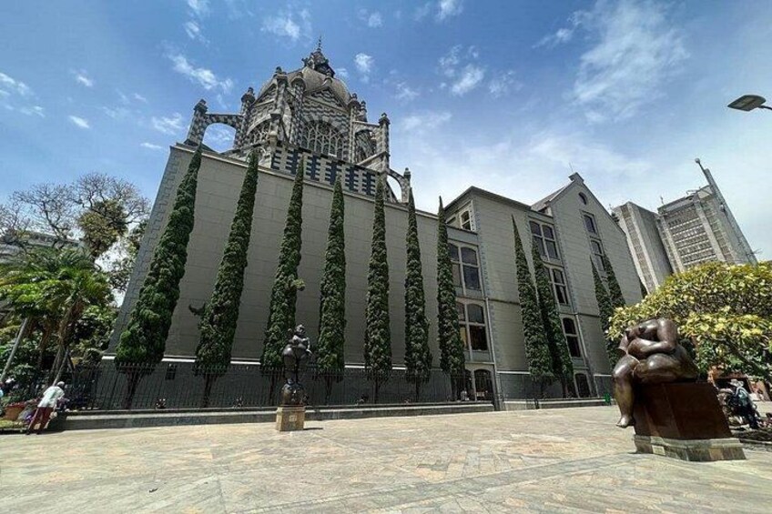 Un edificio grande con una torre distintiva, rodeado por una plaza con una estatua y árboles. 1