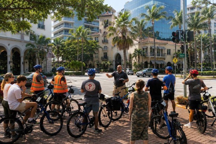 A group of cyclists gathered on a city street with palm trees and buildings in the background. 3