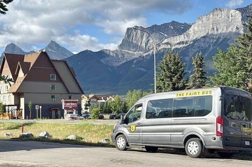 Een zilveren busje, waarop 'The Fairy Bus' staat geschilderd, geparkeerd op een straat met bergen op de achtergrond. 2