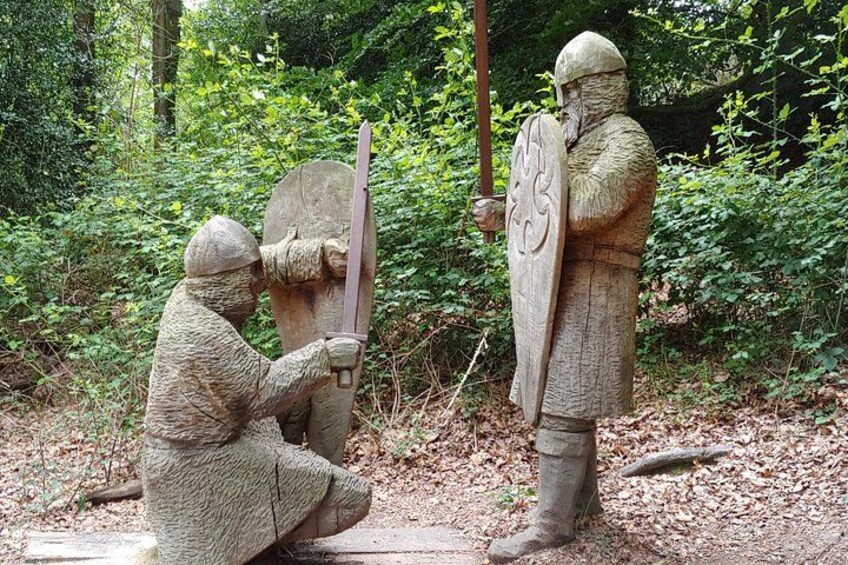 A stone statue of a knight in armor holding a sword and shield, standing in a forested area. 2