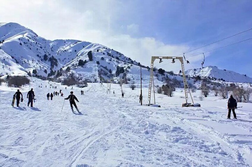 A snowy ski slope with skiers, a ski lift, and mountains in the background. 2