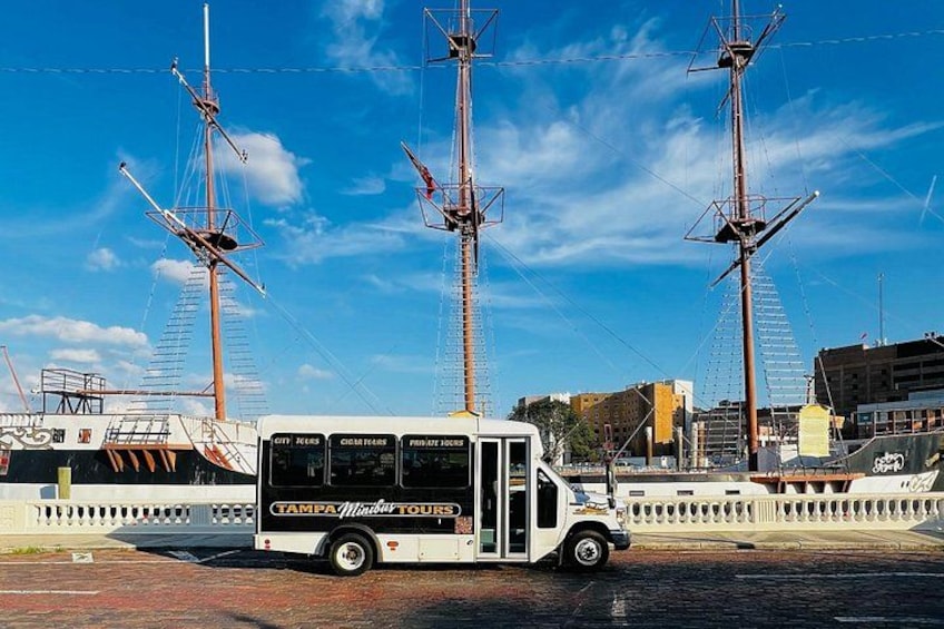 En vit buss med texten ”Tampa Bay Tours” på sidan stod parkerad framför ett stort segelfartyg med tre master. 2