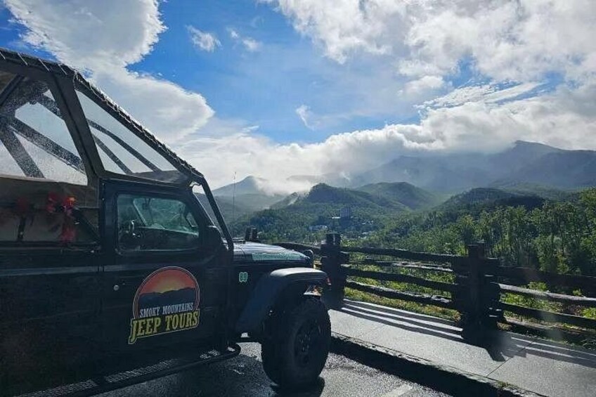 Un SUV nero con la scritta 'JEEP TOURS' sul fianco, parcheggiato su una strada con vista panoramica su montagne e nuvole sullo sfondo. 1