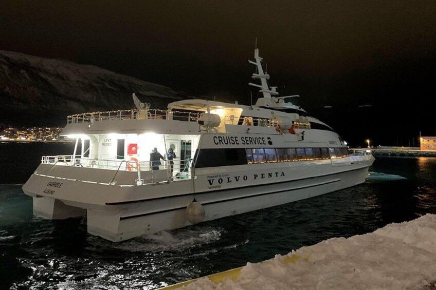 Et krydstogtskib ved navn VOLVO PENTA ligger ved et havneanlæg med et bjergrigt landskab i baggrunden. 2