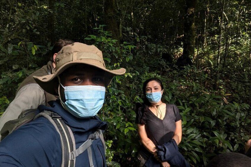 A person wearing a hat and a face mask, another person in a dark shirt, and dense green foliage in the background. 2