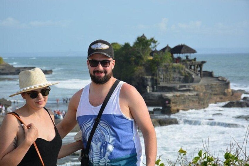 A couple standing by a coastal area with waves crashing onto rocks and a traditional structure in the background. 4