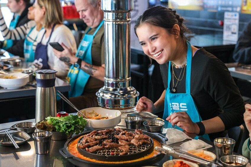 A person wearing a blue apron preparing food at a table with a grill, bowls, and a thermos. 1