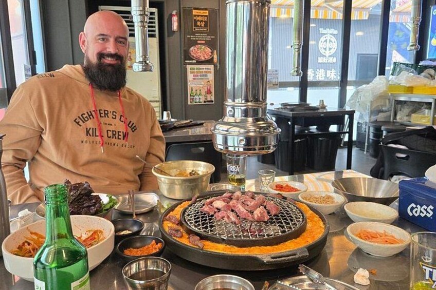 En person sidder ved et bord med en grill, forskellig mad og en flaske grøn væske. 5