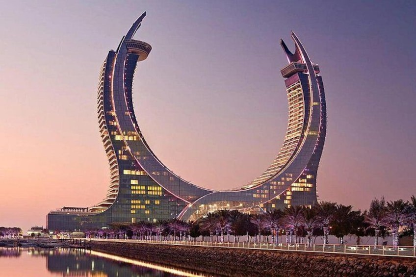 A modern curved building with a reflective facade, surrounded by a waterfront promenade and palm trees. 1