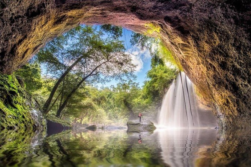 A cave entrance with a waterfall, trees, and a person standing on a rock. 2