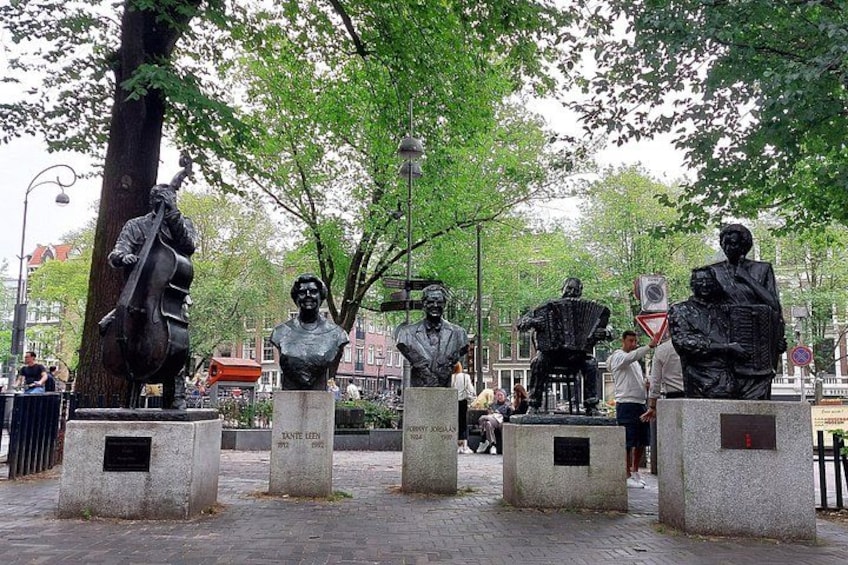 Een park met beelden van muzikanten en bustes op sokkels, omgeven door bomen en een straat. 2
