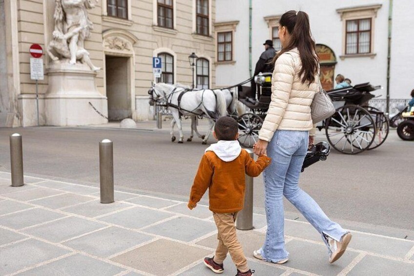 A woman and a child walking on a paved sidewalk, a horse-drawn carriage in the background, and a statue on the left. 3