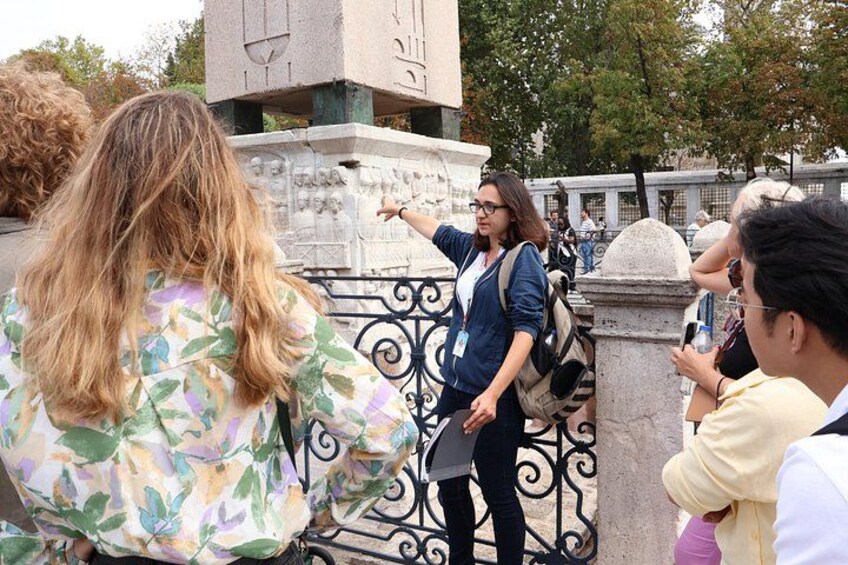 A person pointing at a stone monument with inscriptions, surrounded by a metal fence and trees in the background. 1