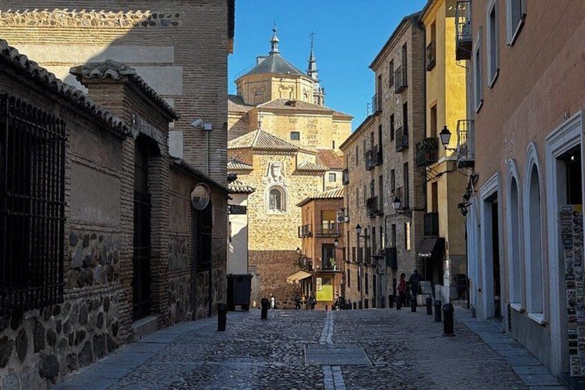 Una calle adoquinada flanqueada por edificios históricos, una iglesia con cúpula y un cielo azul despejado. 1