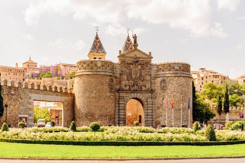 Una puerta de piedra histórica con dos torres, rodeada de un jardín cuidado y edificios urbanos al fondo. 3