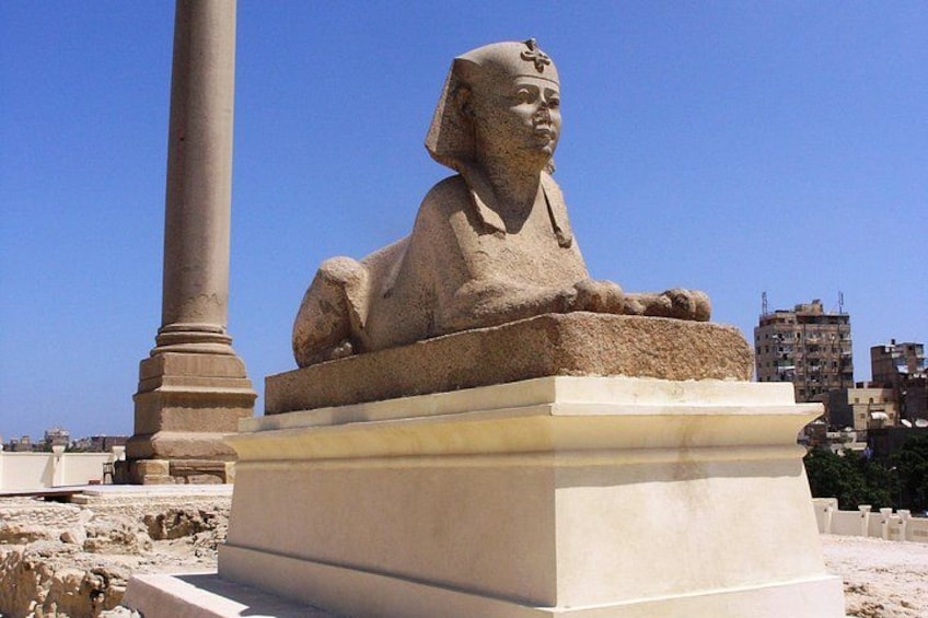 A large stone sphinx statue on a pedestal, with a tall column in the background and modern buildings in the distance. 3