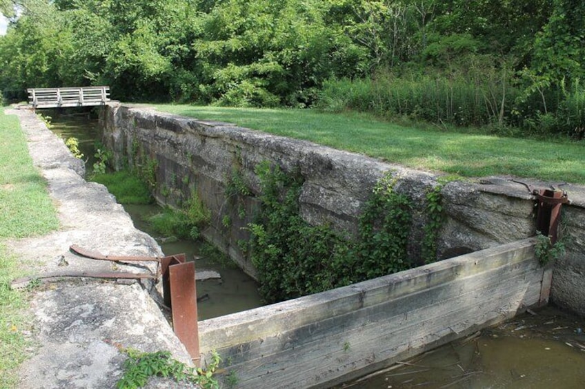 Un estrecho canal de agua con una esclusa de madera, rodeado de vegetación y un pequeño puente al fondo. 5