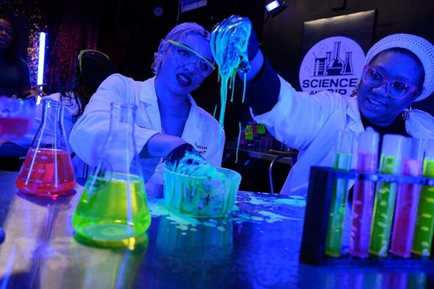 Dos personas con batas de laboratorio frente a un fondo con temática científica, líquidos de colores en vasos de precipitados, y un cartel con la palabra 'SCIENCE'. 4