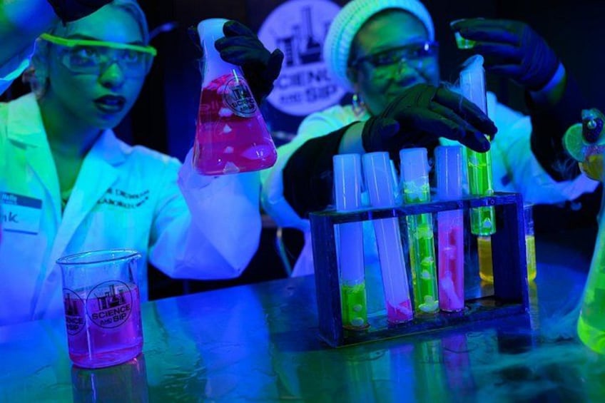 A laboratory setting with people conducting experiments, various colored liquids in test tubes, and a beaker on the table. 2