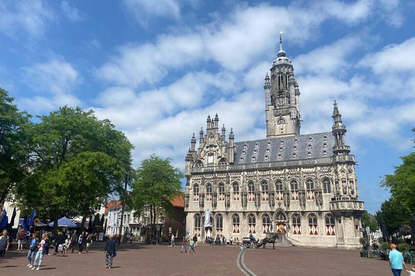 A historic building with intricate architecture, a prominent tower, and a paved square with people and trees. 4