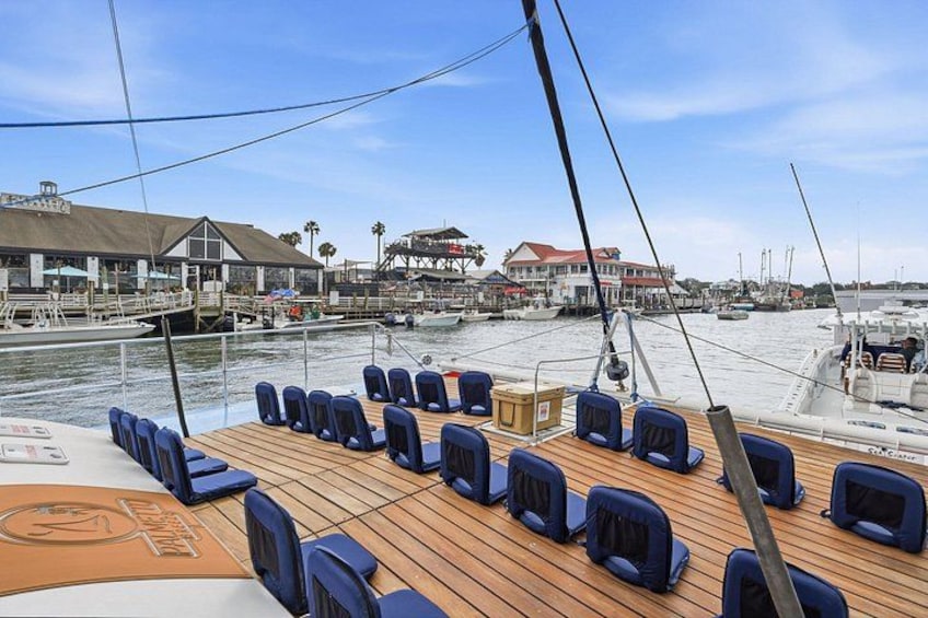 Cubierta de barco con sillas azules, suelo de madera y un puerto deportivo con edificios al fondo. 4