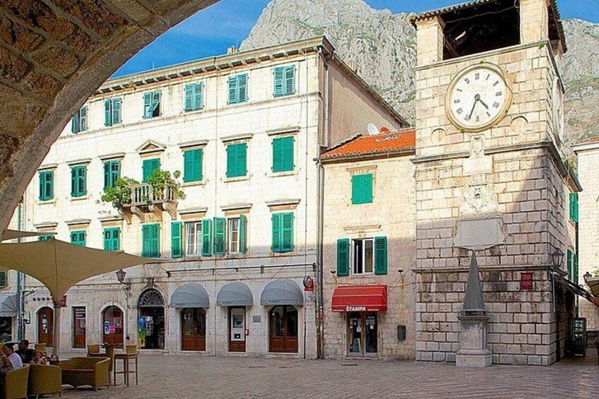 Una plaza histórica con una torre de reloj, edificios con contraventanas verdes y mesas al aire libre. 4