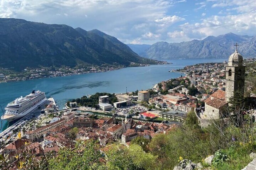 A coastal town with a prominent church, a cruise ship docked, and mountains in the background. 1