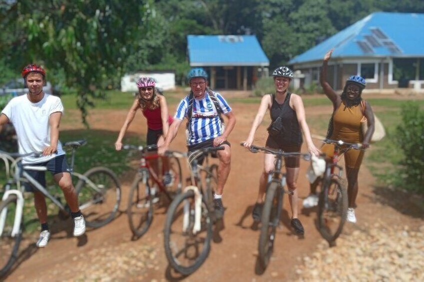 A group of cyclists on a dirt path with trees and buildings in the background. 1