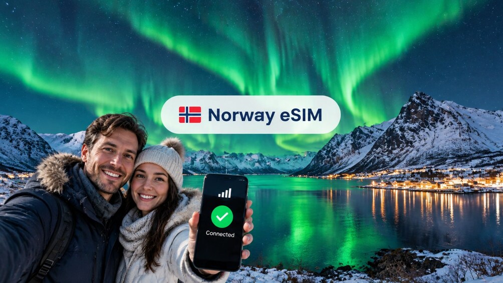 A couple taking a selfie with a smartphone displaying a connected eSIM status, set against a backdrop of the Northern Lights over a snowy landscape with a fjord. 1