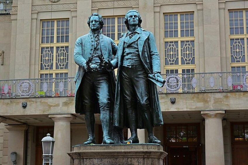 A statue of two figures in period clothing, a building with classical architecture in the background. 1