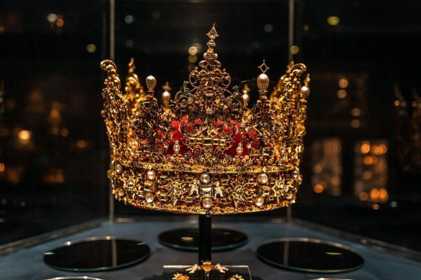 Eine goldene Krone mit aufwendigen Verzierungen und Juwelen, präsentiert auf einem Sockel. 3