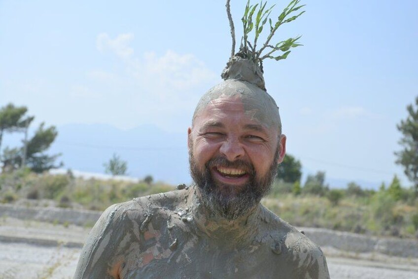 A person covered in mud with a tree branch on their head, standing outdoors with a mountainous background. 4