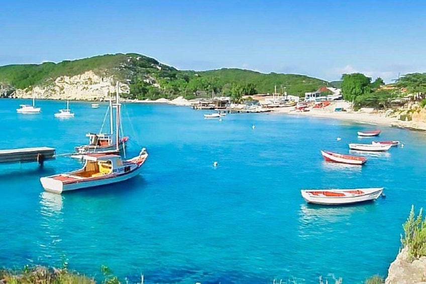 Escena costera con varias embarcaciones ancladas en aguas azules cristalinas, una playa arenosa y un paisaje montañoso al fondo. 3