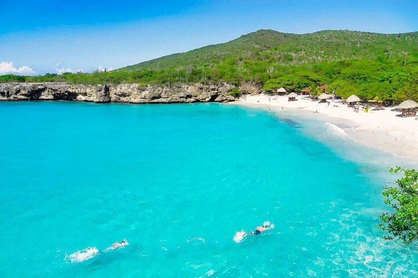 Une plage aux eaux turquoise et limpides, une rive sablonneuse et, au loin, une colline verdoyante. 1