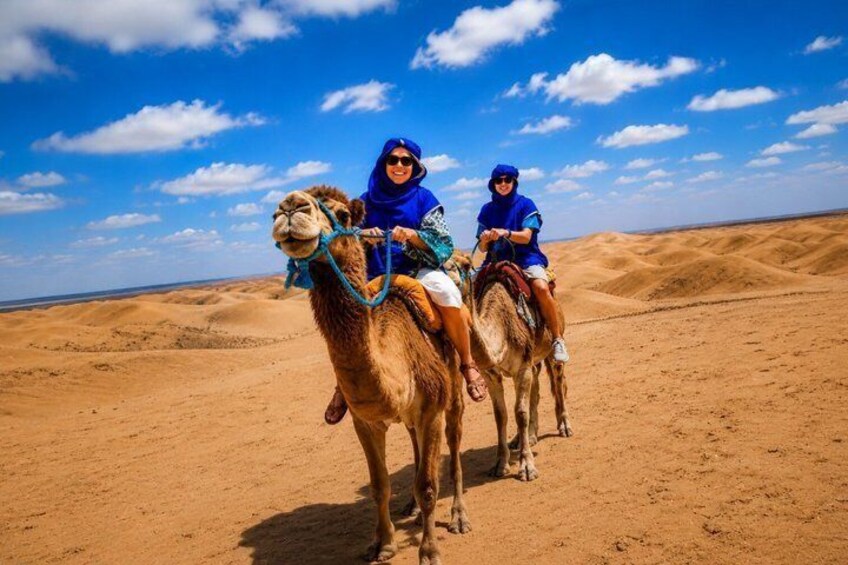 Two people riding camels in a desert landscape with sand dunes and a clear sky. 3