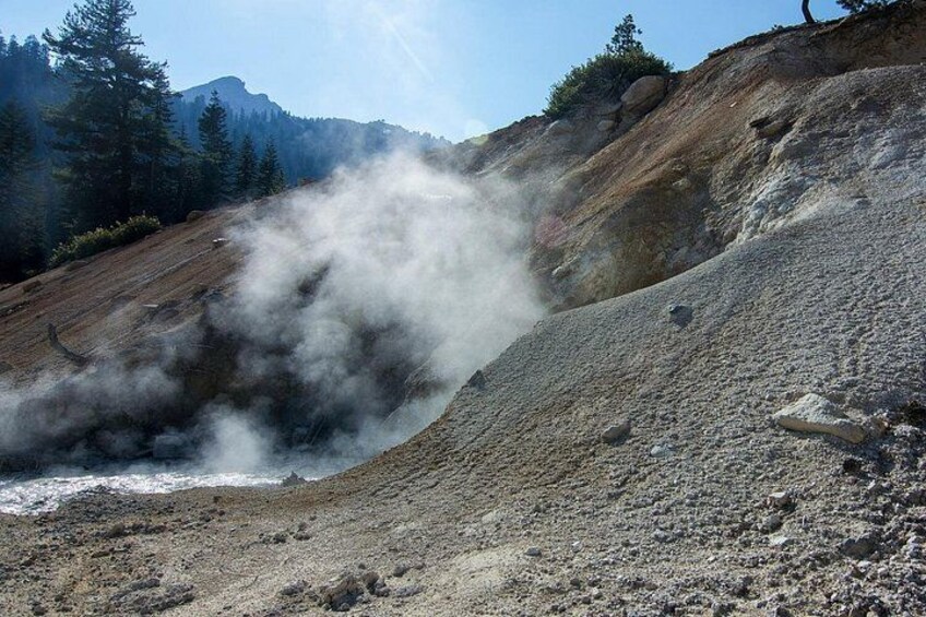 Une zone géothermique où de la vapeur s’échappe du sol, entourée d’un paysage rocheux et d’une végétation clairsemée. 2