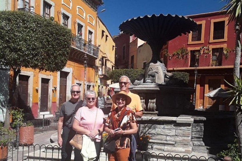 A group of people standing in front of a fountain in a sunny urban setting with colorful buildings. 1