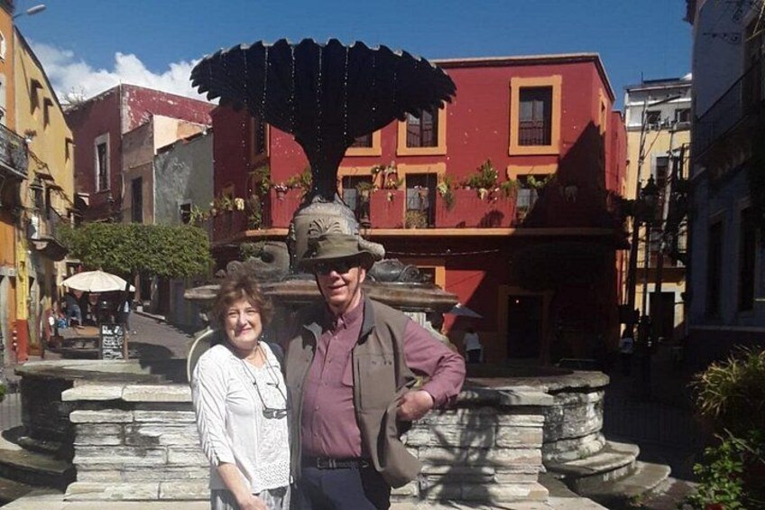 A couple standing in front of a fountain with a statue, surrounded by colorful buildings. 3