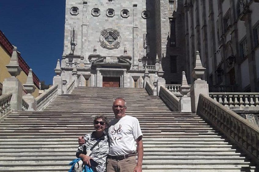 A man and a woman standing on a set of stairs leading to a large building with a clock tower. 4