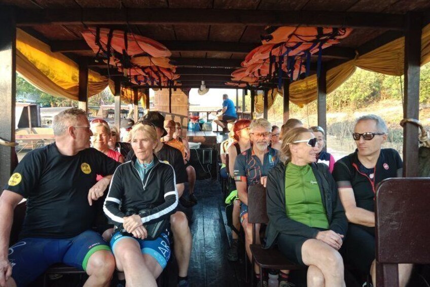 A group of people seated under a covered area with a wooden floor and benches. 2